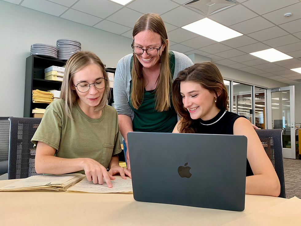 Three women smile and review 19th-century archival papers and a MacBook in an office, working together at a table.