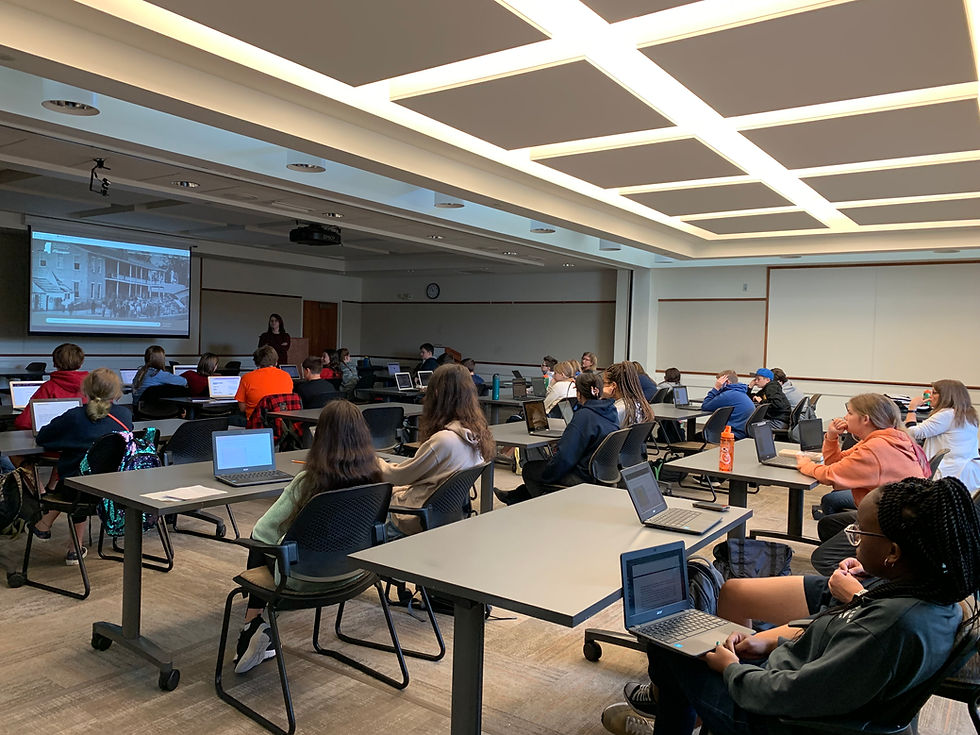 Middle school students sit at laptops in a classroom while a presenter speaks beside a projected slide.