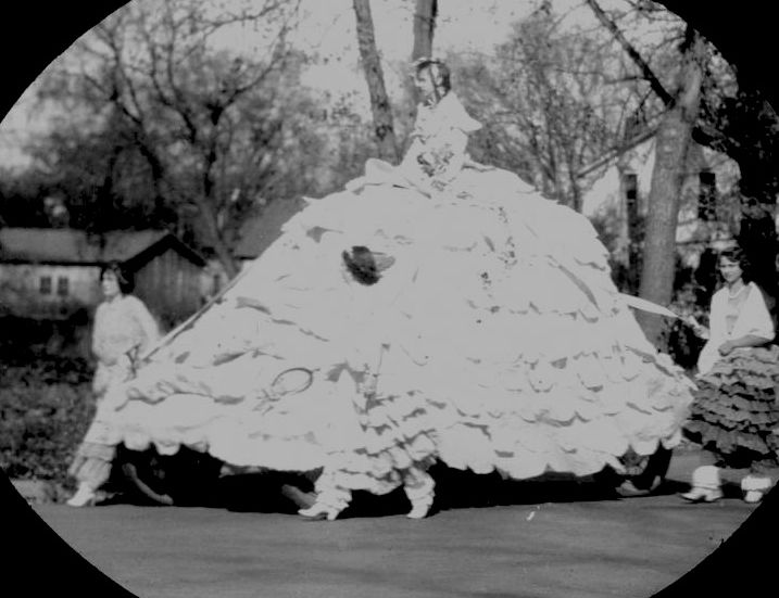 Parade float that is a woman wearing a very frilly and wide hoop skirt.